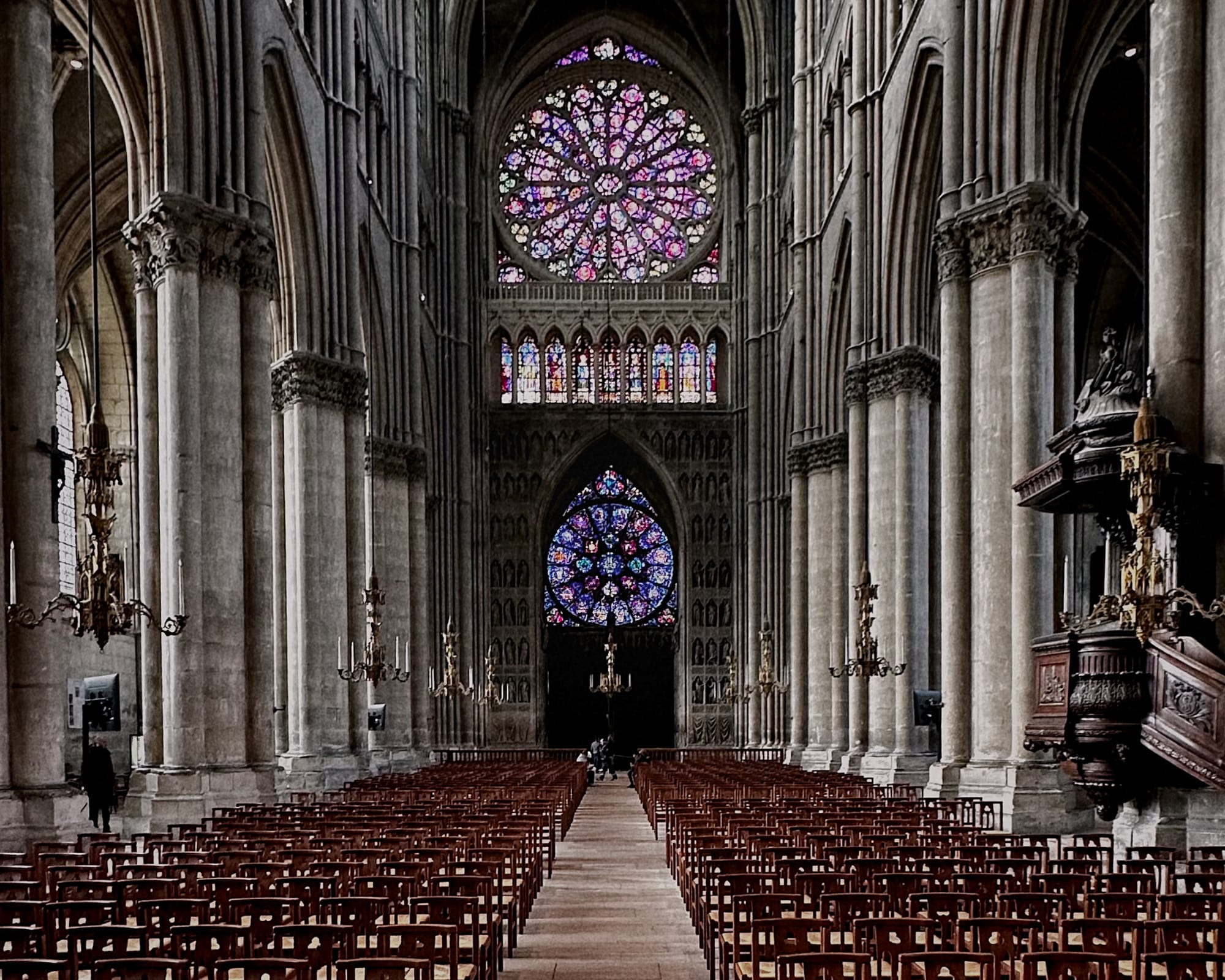 The inside of the Reims Cathedral. Large, round, stained glass windows. Stone pillars. Rows and rows of wooden chairs. 